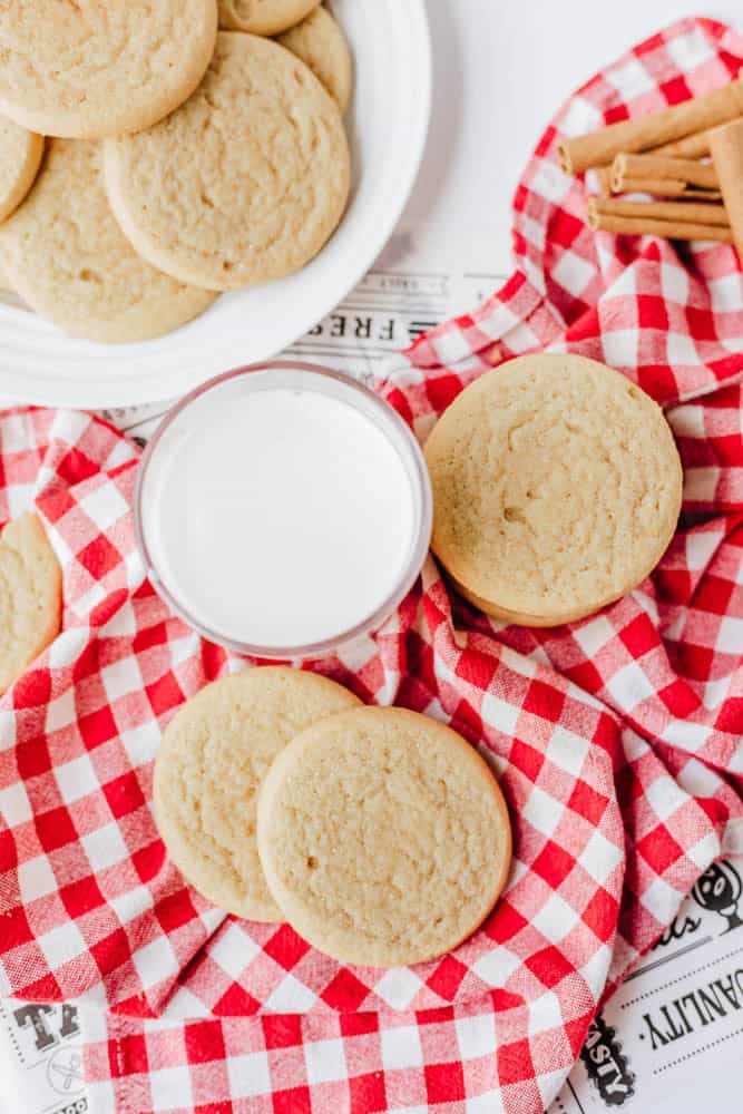 A glass of milk surrounded by several sugar cookies on a red and white checkered cloth, with more cookies on a plate and cinnamon sticks nearby—perfect for enjoying your favorite Snickerdoodle Cookie recipe.