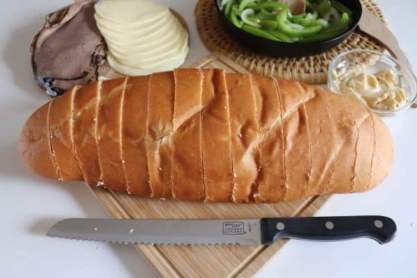 Loaf of french bread cut into 1" slices with philly cheesesteak ingredients on a white table.