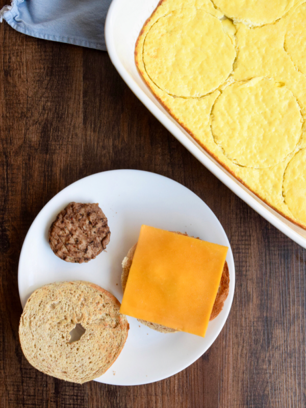 Sliced bagel on a white plate with a slice of cheese and a breakfast sausage.