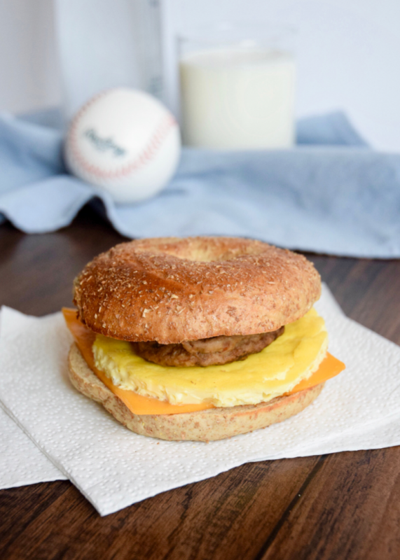 Breakfast sandwich made of a sliced bagel with a sausage pattie, cooked egg and a slice of processed cheese in the middle on a white napkin.