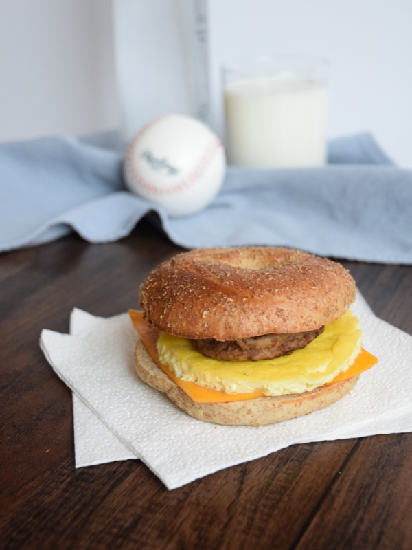 Breakfast sandwich made of a sliced bagel with a sausage pattie, cooked egg and a slice of processed cheese in the middle on a white napkin.