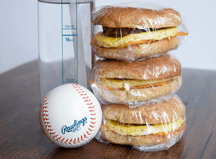 Three camping breakfast bagels wrapped in plastic wrap and stacked on a wooden table.