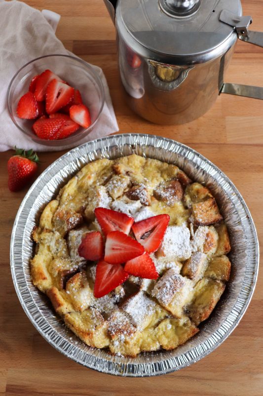 Foil packet french toast bake in a foil pie plate topped with fresh sliced strawberries and powdered sugar.