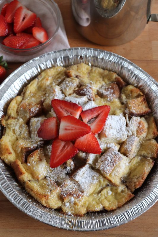 Foil packet french toast bake in a foil pie plate topped with fresh sliced strawberries and powdered sugar.