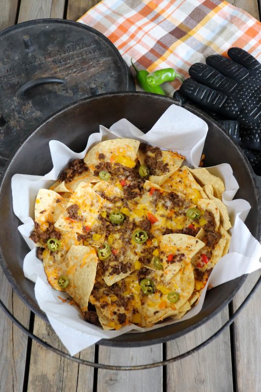 Nachos with ground beef, melted cheese and diced vegetables in a dutch oven lined with parchment paper on a wooden picnic table.