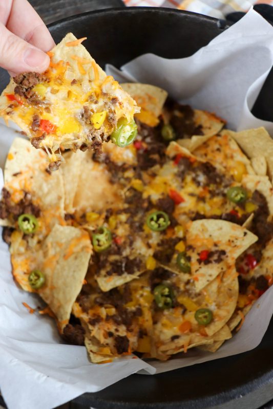 Nachos with ground beef, melted cheese and diced vegetables in a dutch oven with a hand pulling one chip out of the pile.