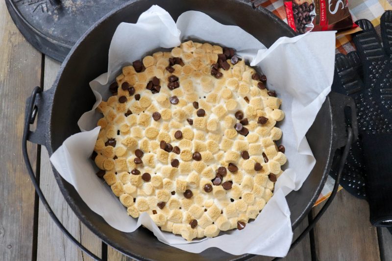 Toasted mini marshmallows and melted chocolate chips in a parchment paper lined cast iron dutch oven on a wooden picnic table.