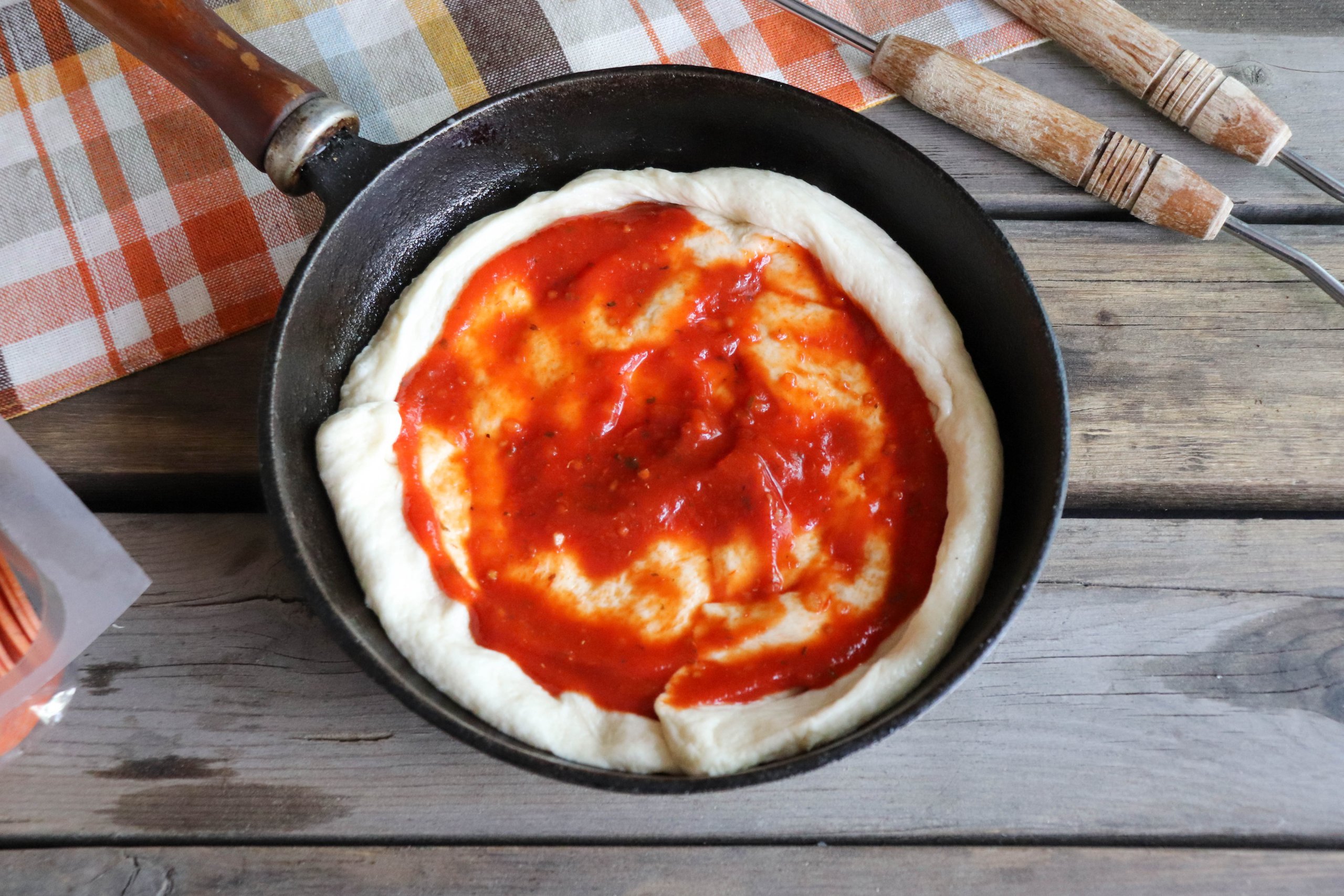 Pizza dough spread onto a cast iron skillet with tomato sauce on a wooden picnic table.