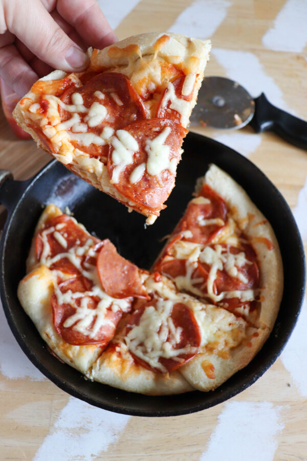 A slice of pepperoni pizza being held in a hand with a skillet full of pizza in the background.