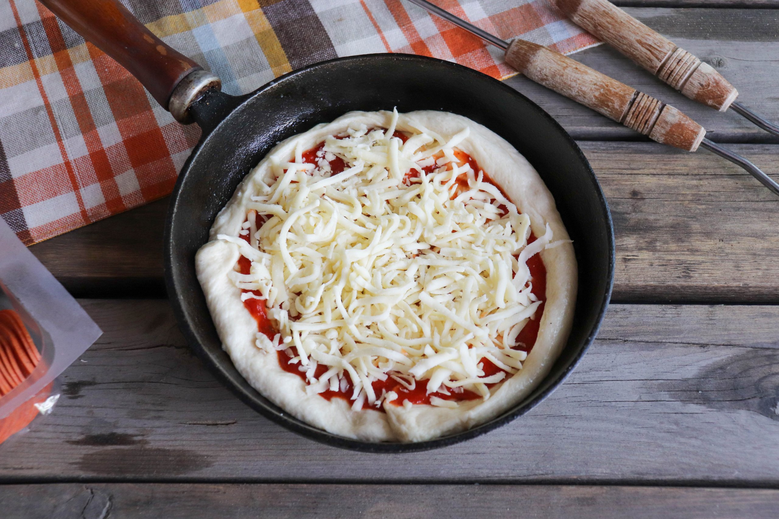 Pizza dough with tomato sauce and shredded mozzarella in a cast iron skillet on a wooden picnic table.