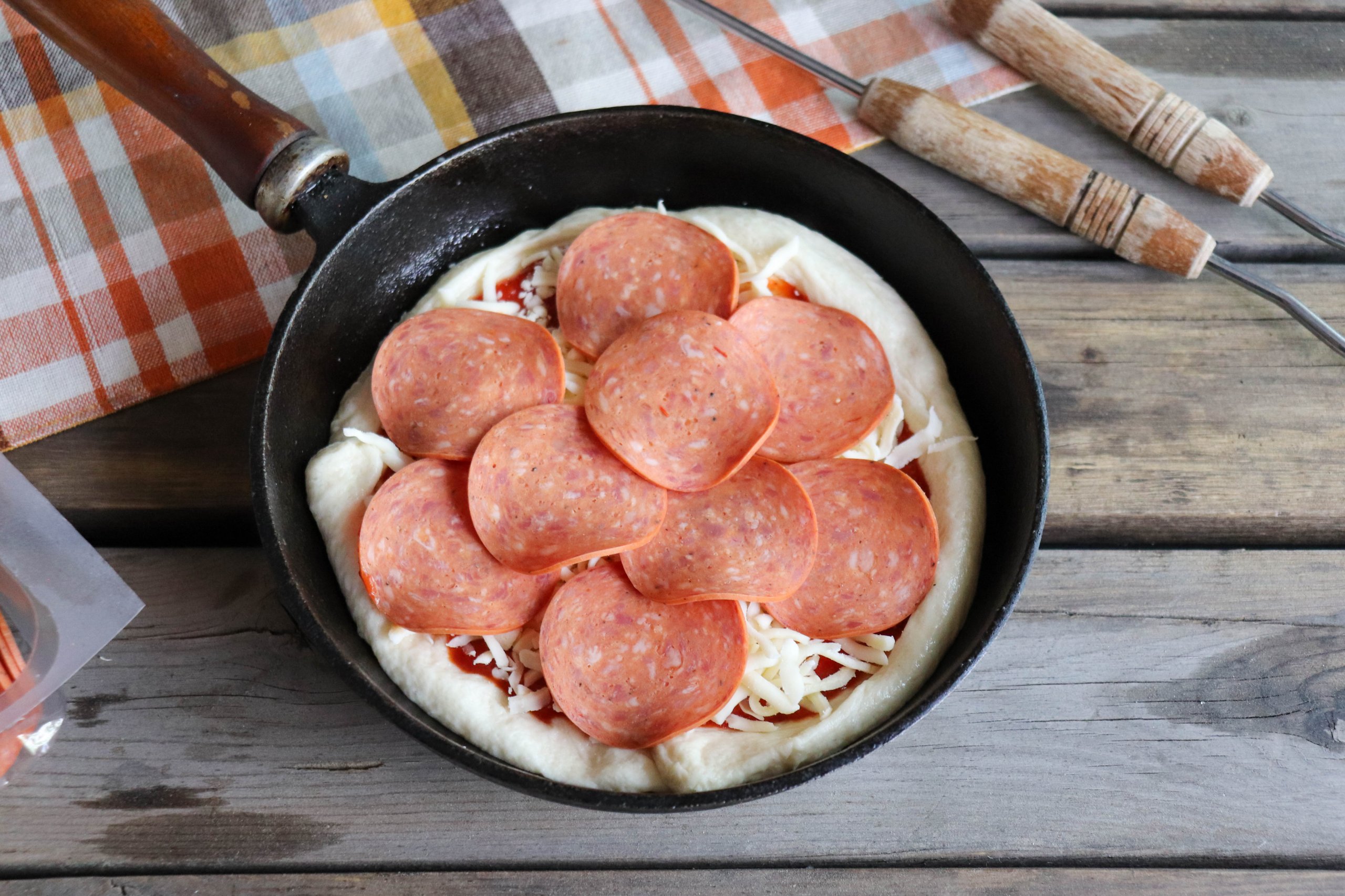 Pizza dough with tomato sauce, shredded mozzarella and pepperoni slices in a cast iron skillet on a wooden picnic table.
