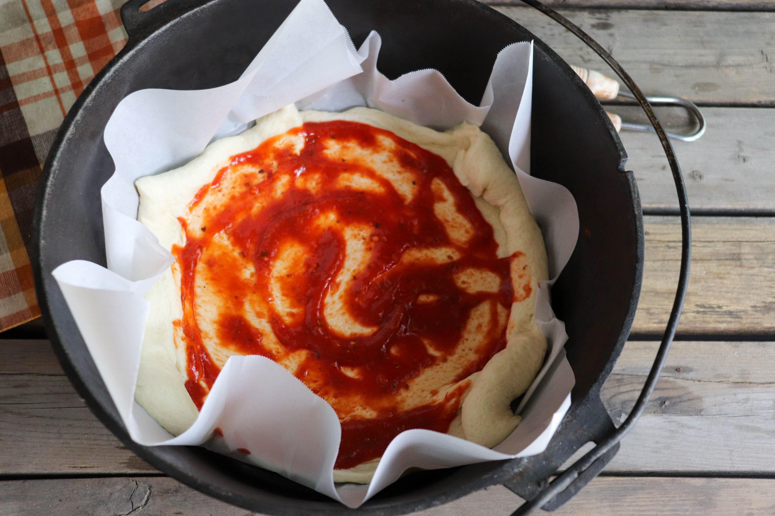 Pizza dough with tomato sauce spread on top in a cast iron dutch oven with parchment paper liner on a picnic table.