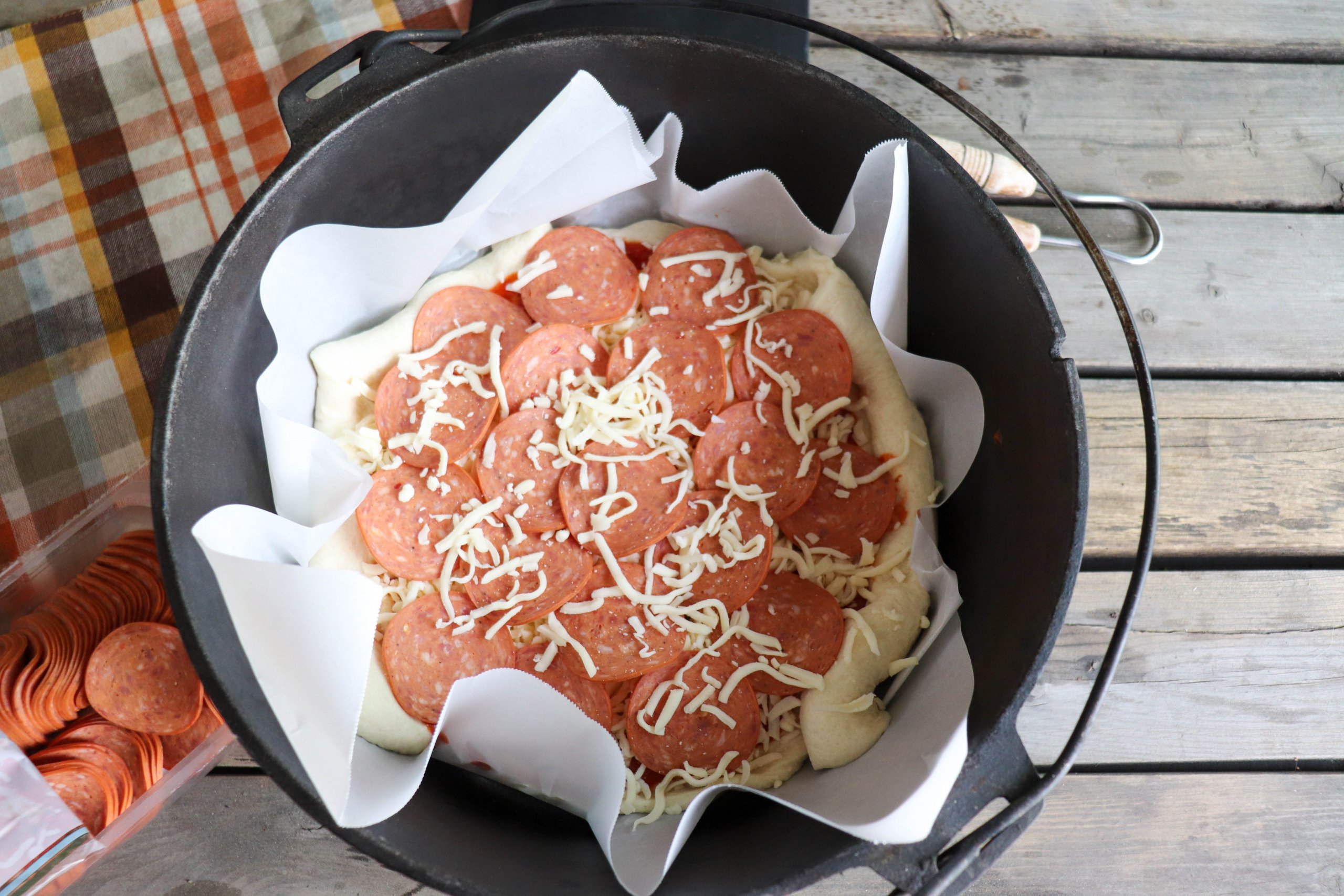 Pizza dough with pepperoni slices and shredded mozzarella cheese on top in a cast iron dutch oven with parchment paper liner on a picnic table.