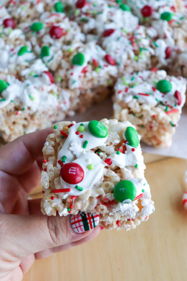 A hand holds a festive Christmas Rice Krispie treat topped with white icing, red and green candy-coated chocolates, and colorful sprinkles. The holiday-themed manicure features a plaid design, with more Christmas Rice Krispie treats in the background.