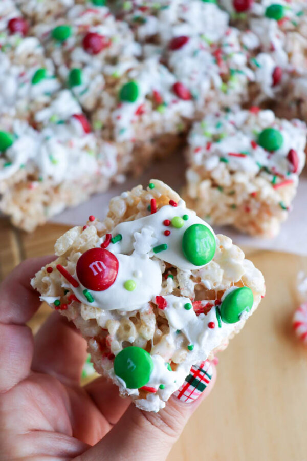 A hand holds a Christmas Rice Krispie Treat topped with white drizzle, red and green M&M candies, and festive holiday sprinkles. More decorated treats are stacked on a wooden surface in the background.