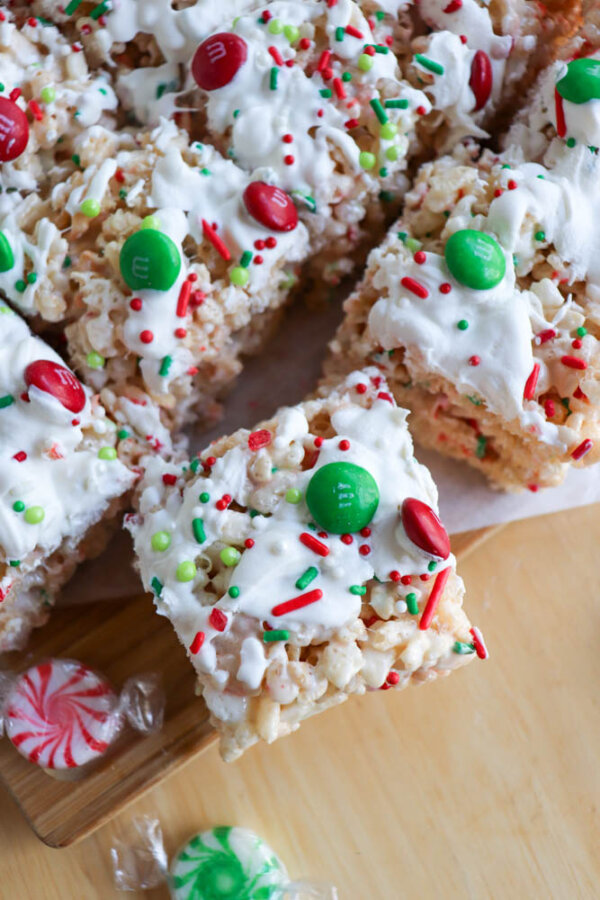 Festive christmas rice krispie treats topped with white icing, red and green M&M candies, and holiday sprinkles are arranged on a wooden board with round peppermint candies nearby.