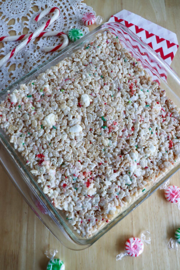 A square glass dish filled with Christmas Rice Krispie treats, topped with white marshmallows and red and green sprinkles, sits on a wooden surface with candy canes and peppermint candies nearby.
