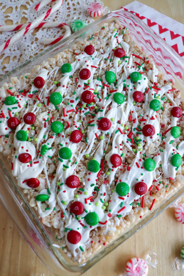 A pan of Christmas Rice Krispie treats topped with white icing, red and green M&Ms, and festive sprinkles sits on a wooden table with candy canes, peppermint candies, and a red-and-white napkin nearby.