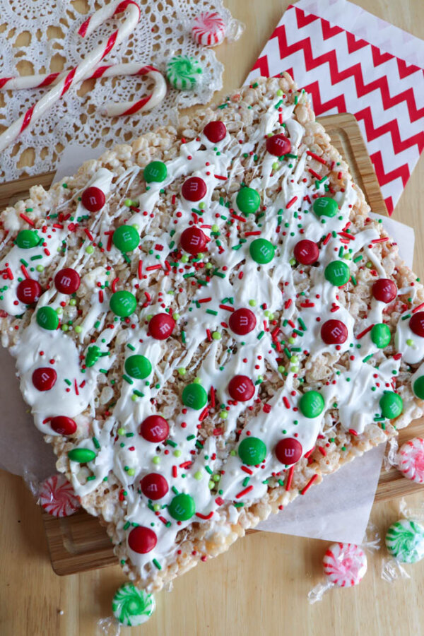 A large, Christmas tree-shaped Rice Krispie treat is decorated with white icing, red and green M&Mโs, and festive sprinkles. Peppermint candies and candy canes are scattered around this fun display of Christmas Rice Krispie treats on a wooden table.