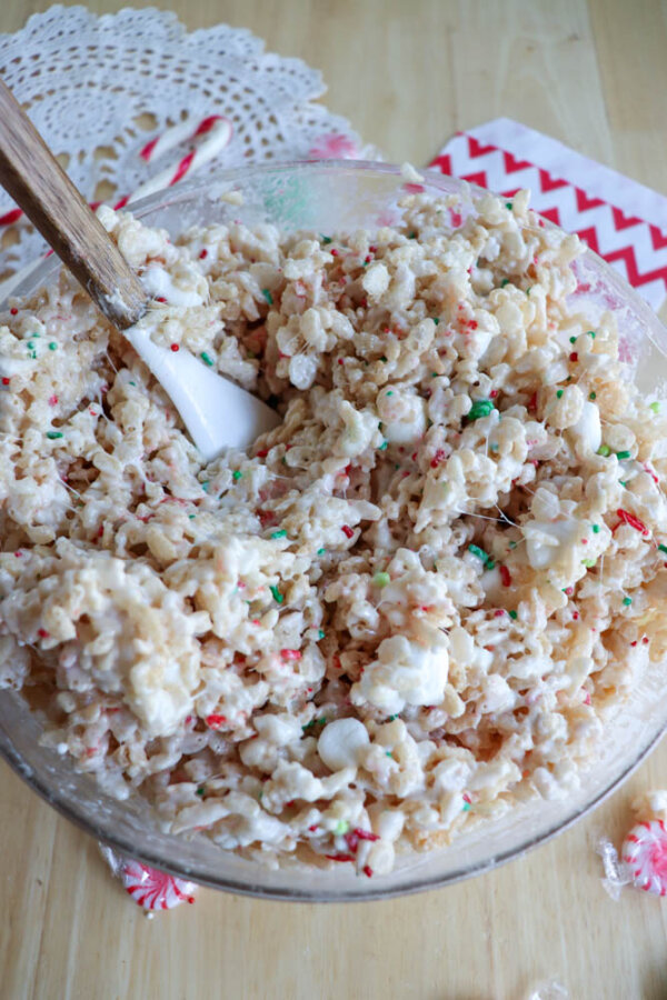 A glass bowl filled with a Christmas Rice Krispie treats mixtureโRice Krispies cereal, melted marshmallows, and red and green sprinklesโis being stirred with a white spatula on a wooden table with festive napkins nearby.