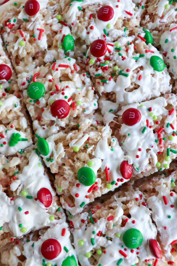 Close-up of festive Christmas Rice Krispie treats topped with white icing, red and green M&M candies, and colorful holiday sprinkles, cut into squares and arranged closely together.