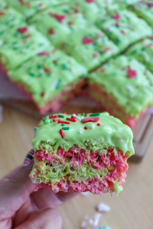A close-up of a hand holding a bright, colorful Grinch Rice Krispie Treat with green frosting and red and green sprinkles. More treats are visible in the background, cut into festive squares.