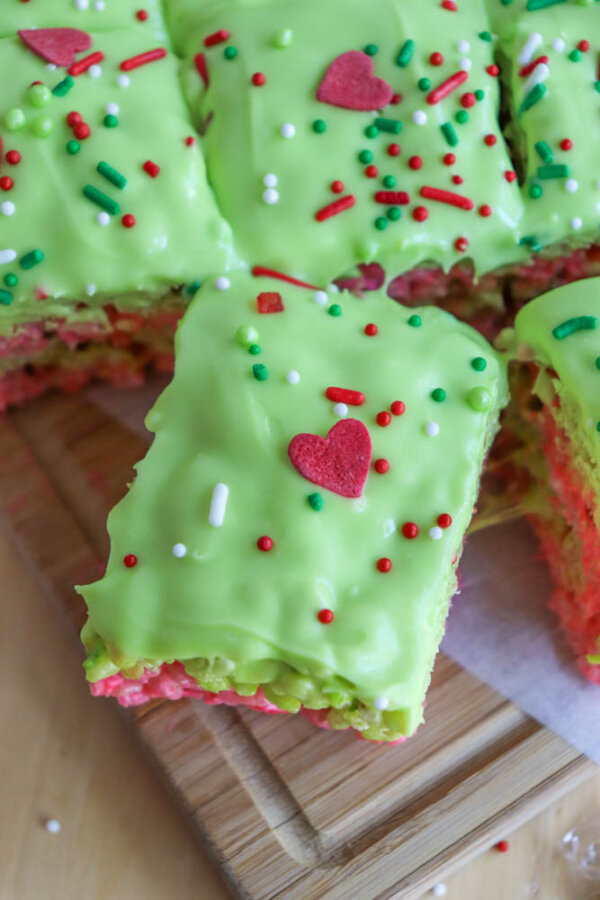 A close-up of Grinch Rice Krispie Treats: bright green-frosted squares topped with red heart-shaped sprinkles and colorful nonpareils, cut into pieces on a wooden board.