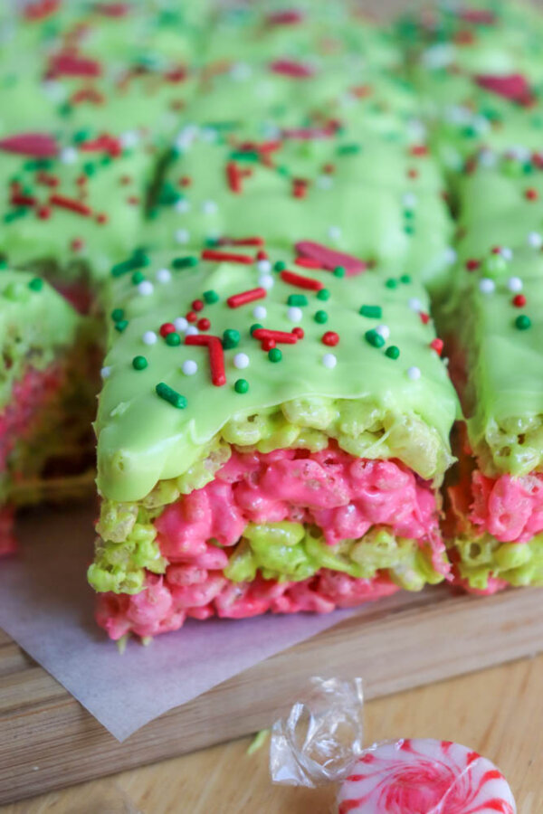 A close-up of Grinch Rice Krispie Treats layered in pink and green, topped with green icing and red, white, and green sprinkles, arranged on a wooden surface with a mint candy nearby.