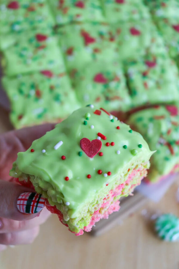 A hand with plaid-patterned nails holds a festive Grinch Rice Krispie Treat topped with green frosting, red, white, and green sprinkles, and a small red heart. More decorated treats are visible in the background.