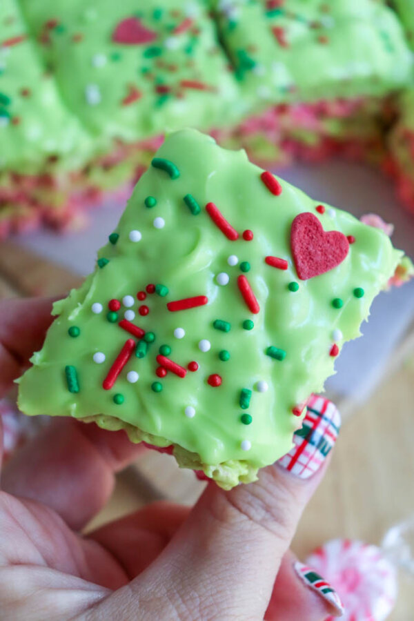 A hand holding a festive Grinch Rice Krispie Treat topped with red, white, and green sprinkles and a red heart, with more treats in the background. The person has plaid-patterned holiday nail art.