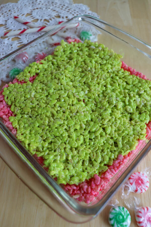 A glass baking dish filled with Grinch Rice Krispie Treats in pink and green sits on a wooden surface, accented by peppermint candies and a lace doily nearby.