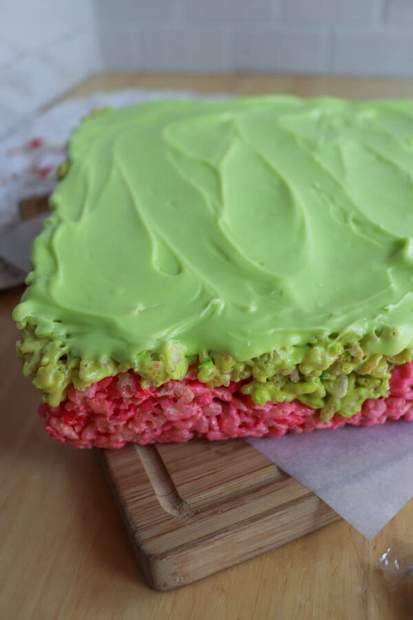A rectangular Grinch Rice Krispie Treat with a pink bottom layer, green middle, and smooth light green frosting on top, sits on a wooden cutting board.