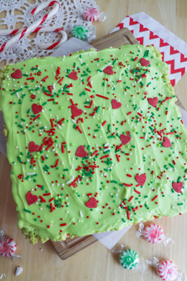 A square Grinch Rice Krispie Treat with light green frosting, topped with red, white, and green sprinkles and heart-shaped decorations, sits on parchment paper on a wooden surface, surrounded by peppermint candies and festive decor.