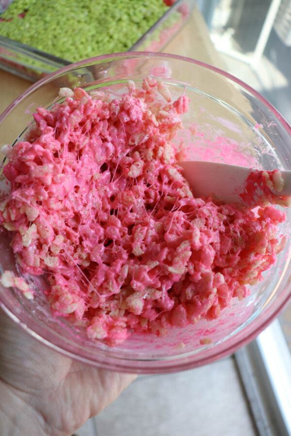 A hand holds a glass bowl filled with pink, gooey Grinch Rice Krispie Treat mixture being stirred with a white spatula; a green tray of Grinch Rice Krispie Treats sits in the background.