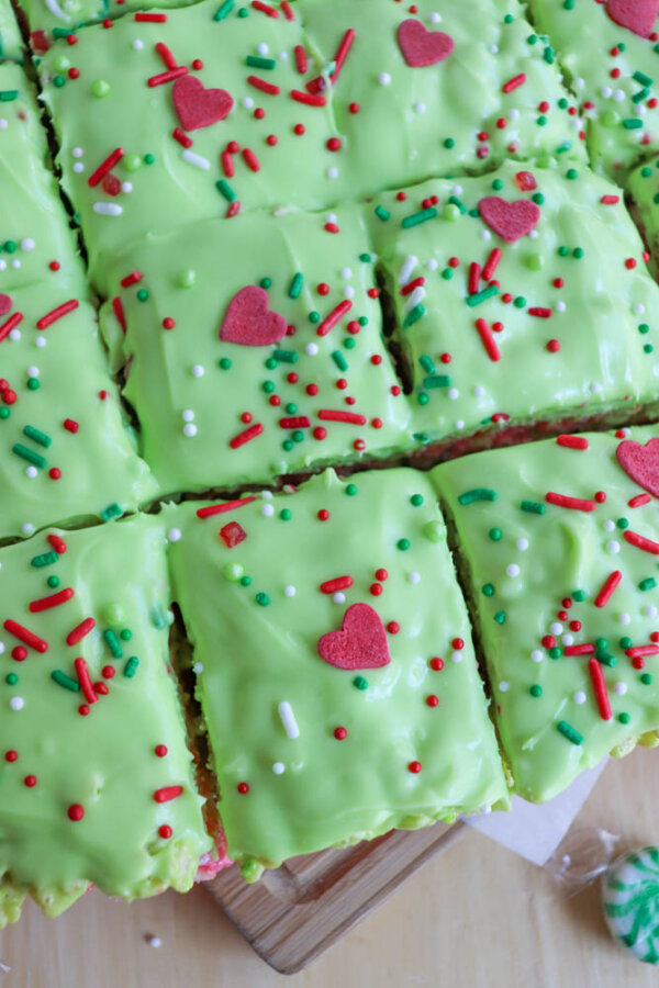 Grinch Rice Krispie Treats: Sliced green-frosted dessert bars topped with red, white, and green sprinkles, plus red heart-shaped decorations, arranged on a wooden surface.