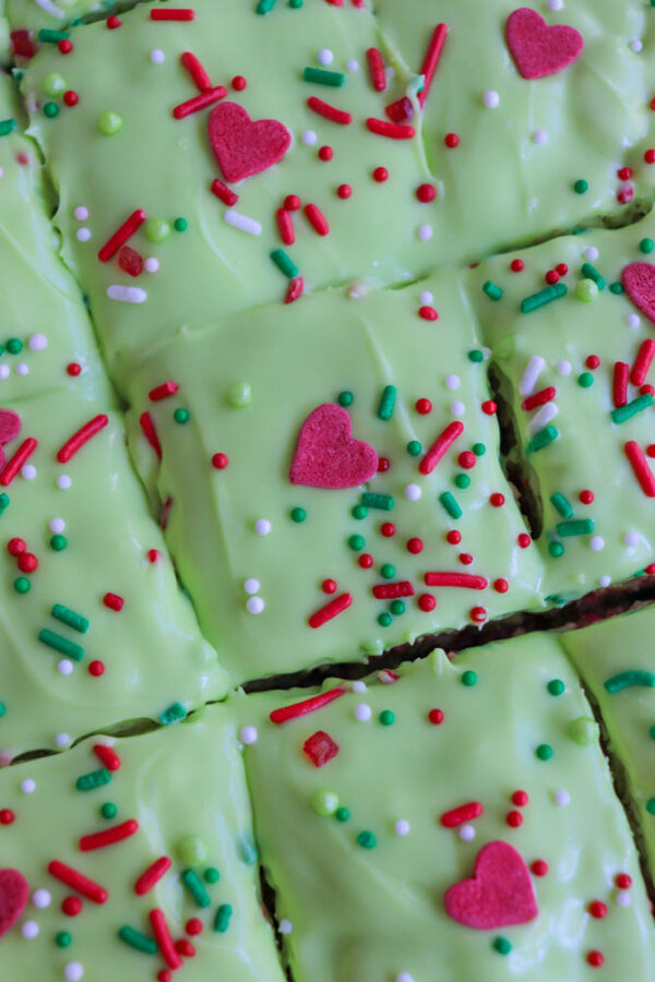 Close-up of Grinch Rice Krispie Treats—green frosted cake squares topped with red heart-shaped sprinkles, plus red, white, and green round and rod-shaped sprinkles, all arranged in a festive grid pattern.
