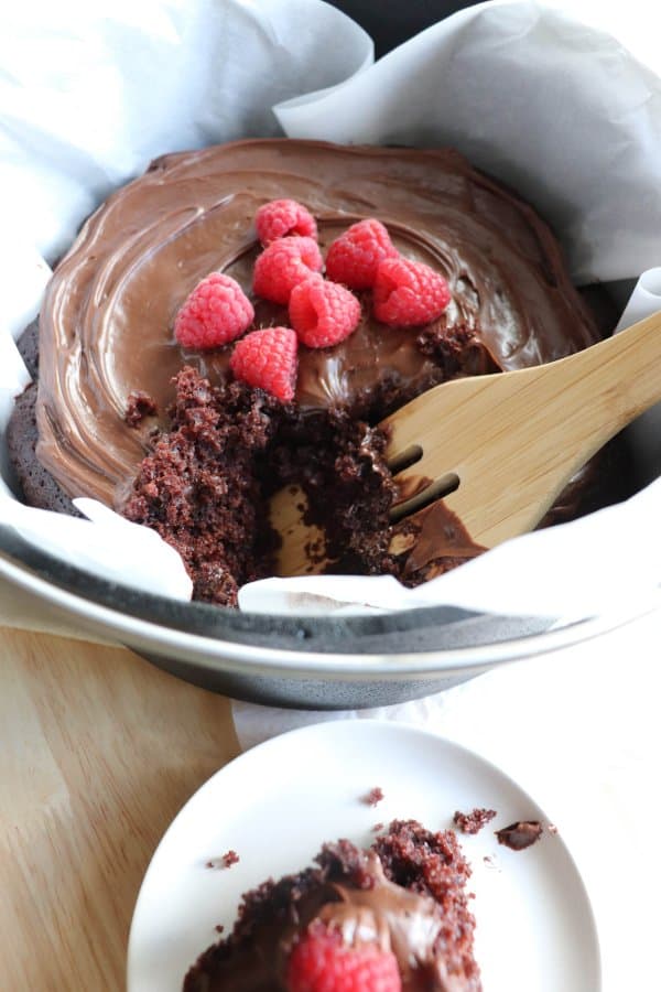 Chocolate Cake with chocolate frosting topped with fresh raspberries in a parchment paper lined cast iron dutch oven with a scoop taken out and put on a plate.