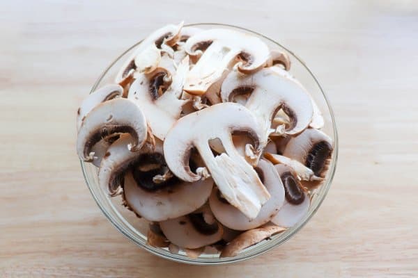 skillet philly cheesesteak recipe Raw sliced Mushrooms in a glass bowl on a wooden table.