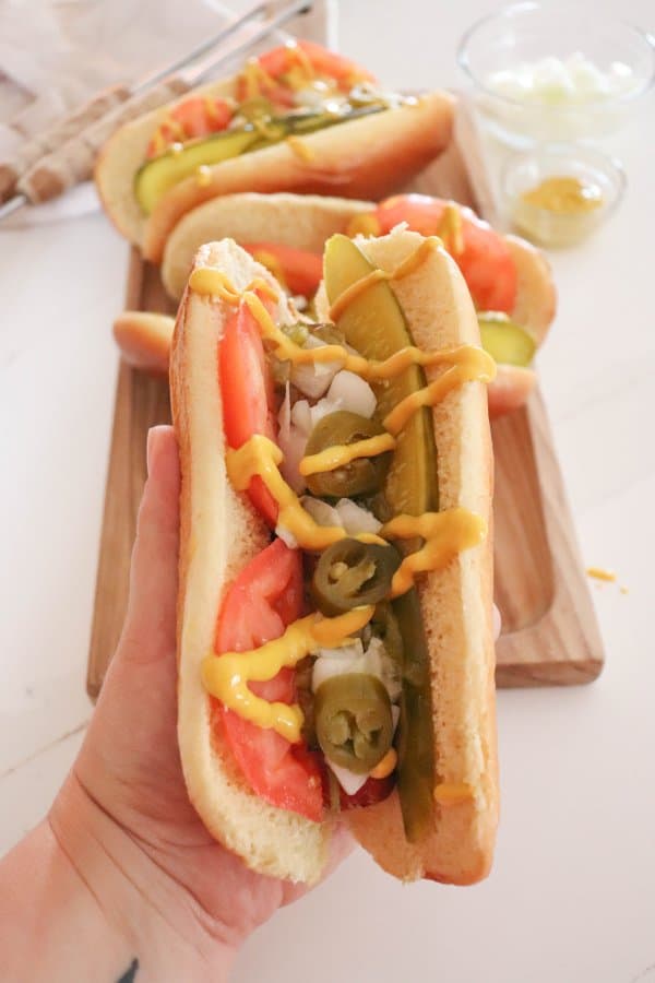 Chicago hot dog topped with pickle spear, sliced tomato, diced onion, pickled peppers and mustard held in a hand.
