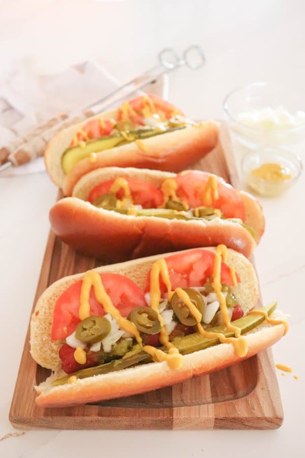 Three Chicago hot dogs topped with pickle spears, sliced tomatoes, diced onion, pickled peppers and mustard on a wooden cutting board.