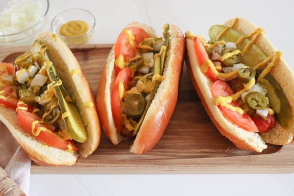 Three Chicago hot dogs topped with pickle spears, sliced tomatoes, diced onion, pickled peppers and mustard on a wooden cutting board.