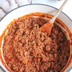 A white pot filled with cooked minced meat in a rich, red hot dog sauce recipe is being stirred with a wooden spoon. A white cloth and a pot lid are in the background on a light wood surface.