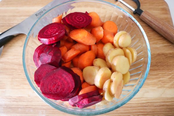 Cut up beets, carrots and potatoes in a clear glass bowl on a wooden surface.