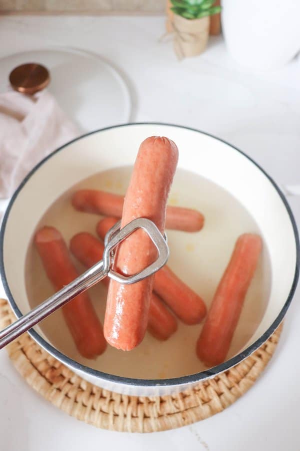 Boiled hot dog held by tongs over a pot of hot dogs.