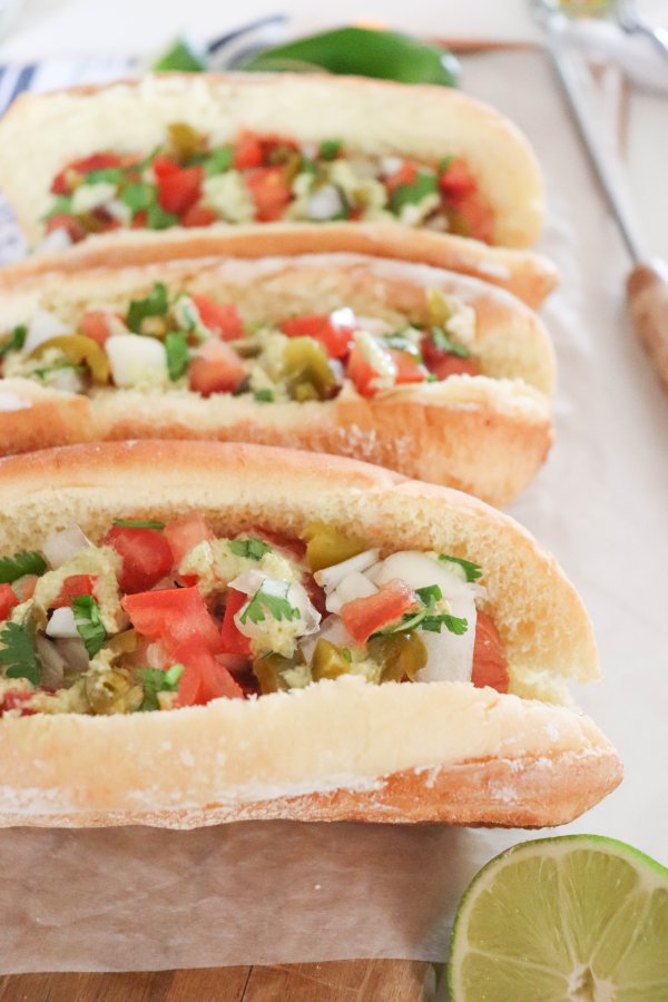 Three open sandwich buns filled with diced tomatoes, onions, cilantro, and green peppers are lined up on parchment paper, reminiscent of a classic mexican hot dog recipe, with a lime half and a knife visible nearby.