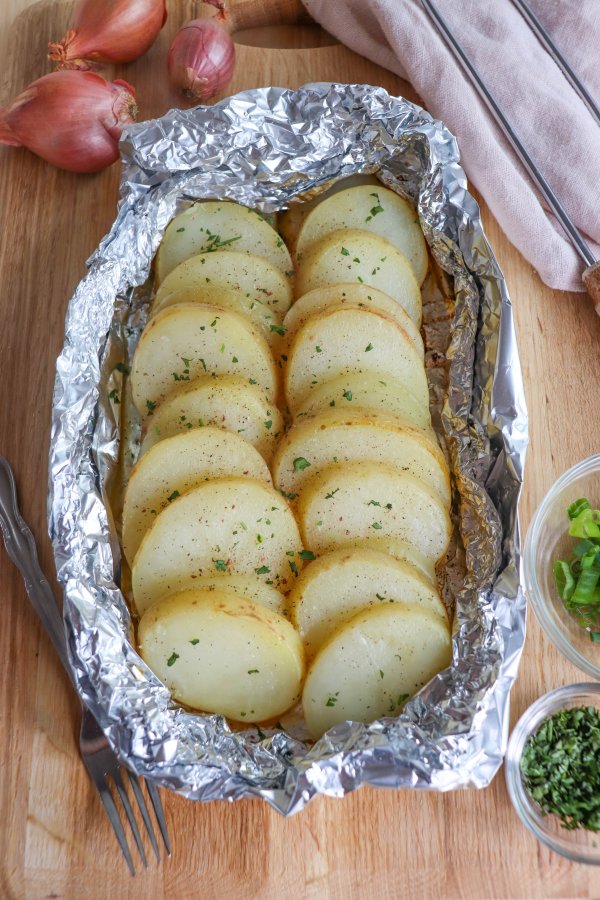 Sliced and seasoned potato and onion in foil packs on a wooden surface.