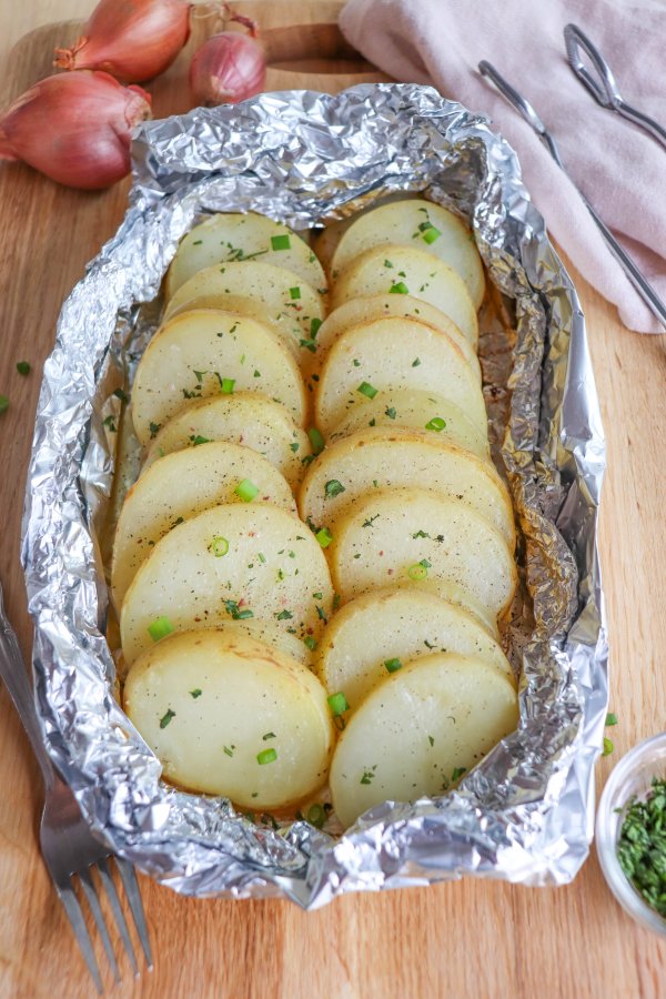 Sliced and seasoned potato and onion in foil packs on a wooden surface.
