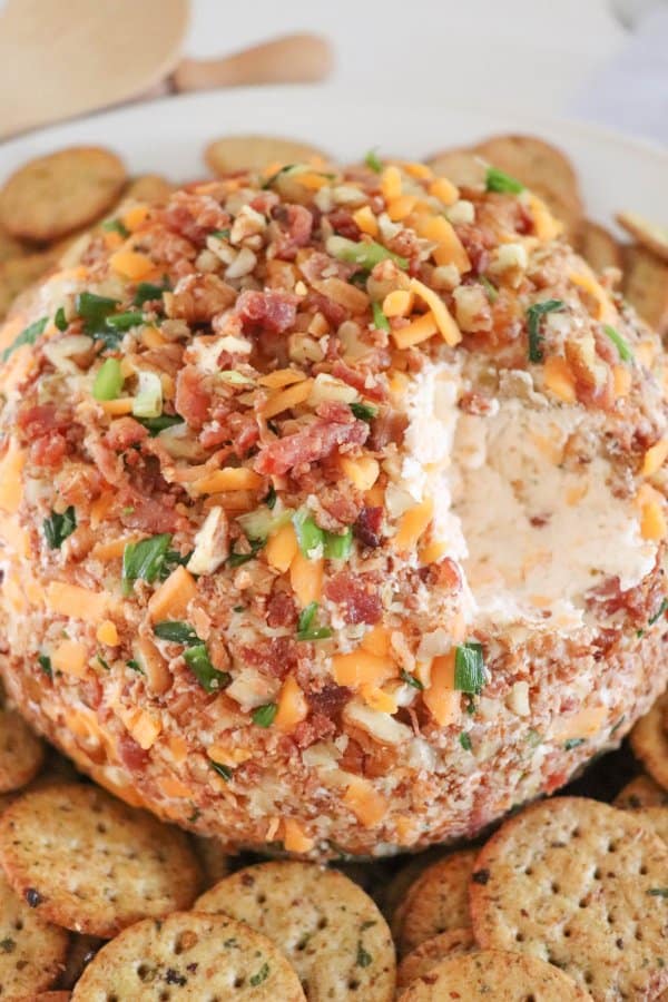 A delicious bacon ranch cheese ball recipe is covered in chopped nuts, bacon, and herbs, perfectly placed on a white plate. Surrounding it are several round crackers arranged in a circle. A portion of the cheese ball has already been enticingly scooped out.