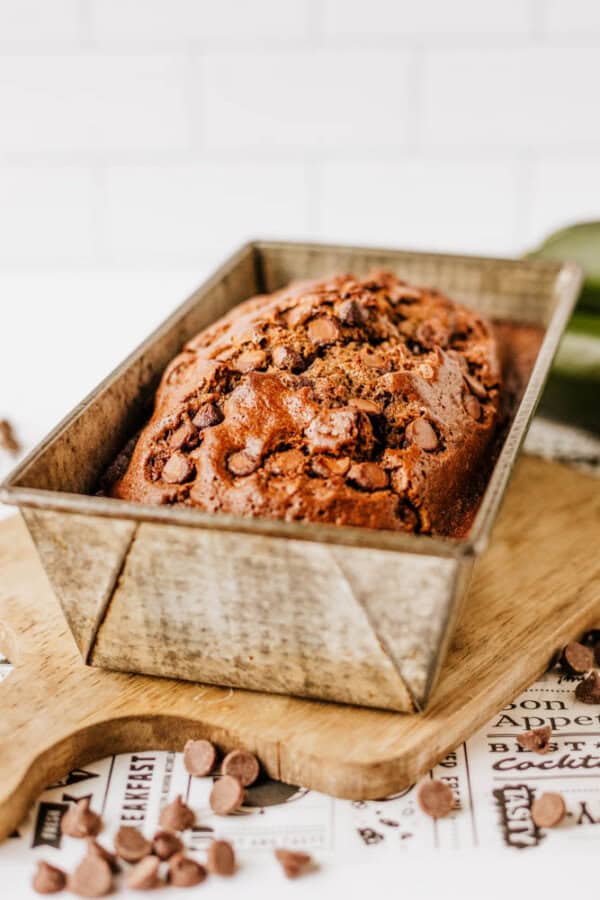 A loaf of chocolate chip banana bread sits in a metal baking pan on a wooden cutting board, with chocolate chips scattered around—a tempting treat for any chocolate zucchini bread lover—against a white tiled background.
