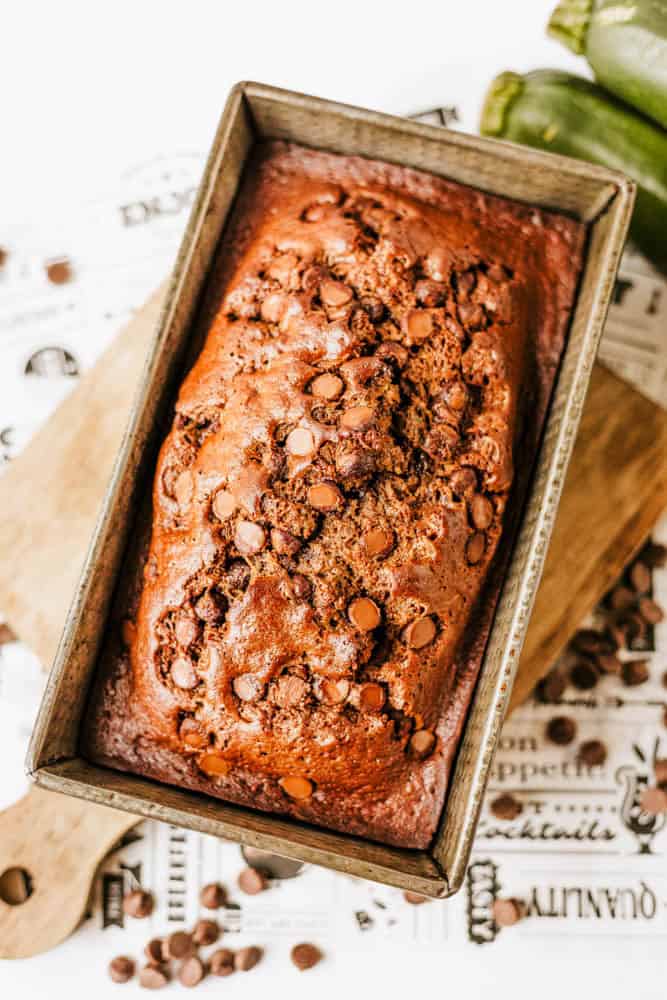 A loaf of chocolate zucchini bread in a rectangular metal pan, topped with melted chocolate chips and resting on a wooden cutting board. Chocolate chips are scattered nearby.
